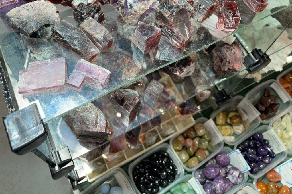 Витрина с минералами и полудрагоценными камнями на ежегодной выставке «Гемма» в Москве. На стеклянных полках представлены необработанные образцы красноватых и фиолетовых минералов, среди которых могут быть эвдиалит, лепидолит или другие редкие породы. Внизу видны контейнеры с полированными камнями различных цветов, включая фиолетовые, черные, зеленые и оранжевые образцы