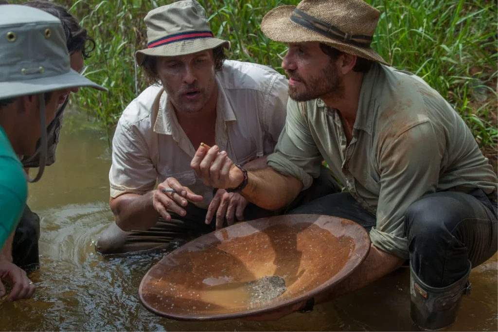 Группа мужчин в походной одежде и шляпах стоит в мелкой реке, держа деревянный лоток с намытой породой, вероятно, содержащей золото.