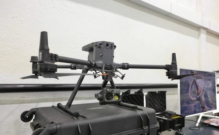 Квадрокоптер с камерой и сенсорами, установленный на черный кейс с колесами, в помещении с белыми стенами. На заднем плане виден монитор с изображением и другие аксессуары для дрона