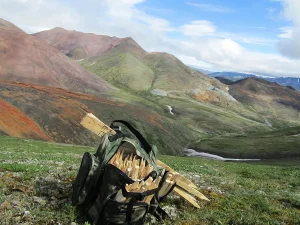 Рюкзак, наполненный пикетами для разметки профиля, стоит на склоне горы. Вдали видны разноцветные горные породы и разработка карьера на золото