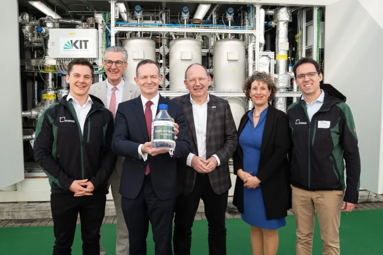 Группа людей в деловой и рабочей одежде стоит перед технологической установкой KIT, держа бутылку с надписью 'Зеленый метанол'