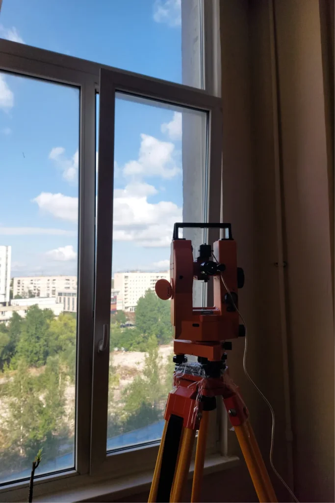Геодезическое оборудование у окна с видом на городскую застройку и голубое небо