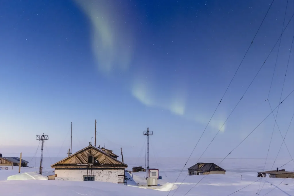 Северное сияние над полярной геофизической станцией с антеннами и деревянными домами на фоне звёздного неба