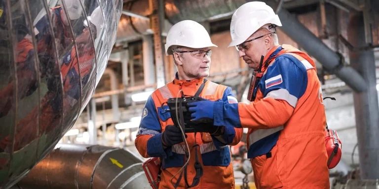 Два инженера в защитных касках и спецодежде обсуждают данные на планшете в промышленном цехе