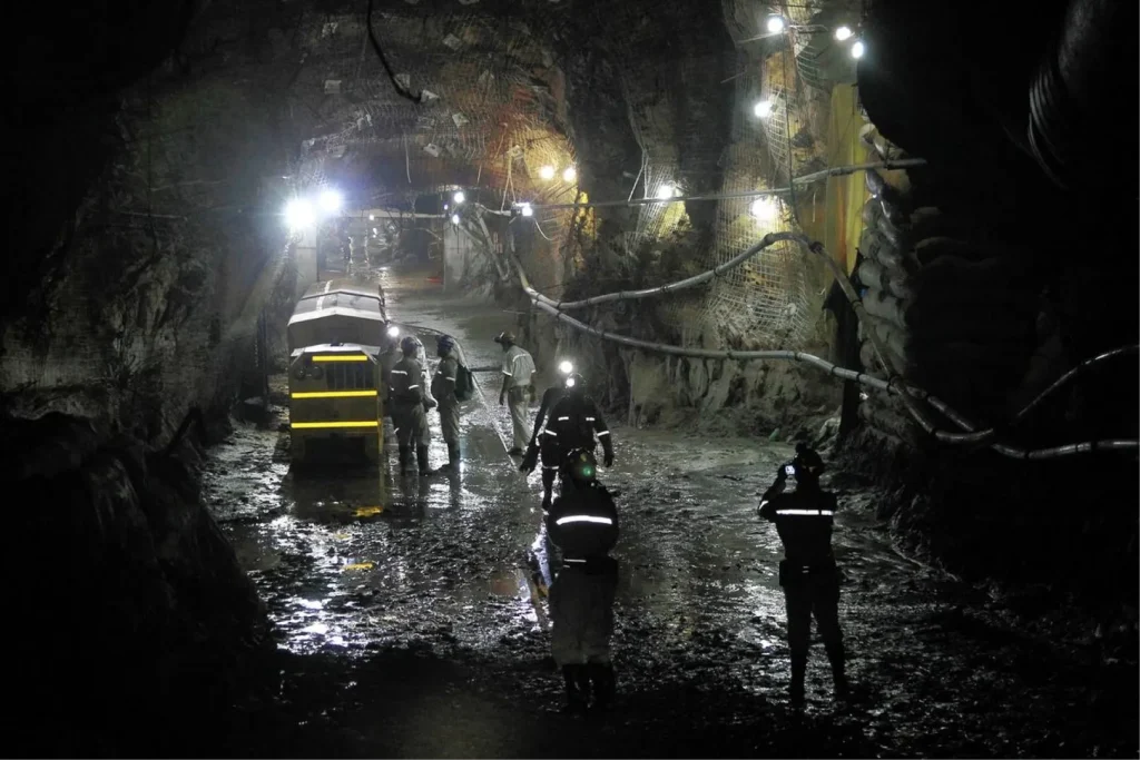 Горняки и техника в тоннеле шахты Mponeng — глубинного рудника, где золото добывают на уровне более 4 км под землёй. Освещённый рабочий участок с дренажом и инженерными коммуникациями.