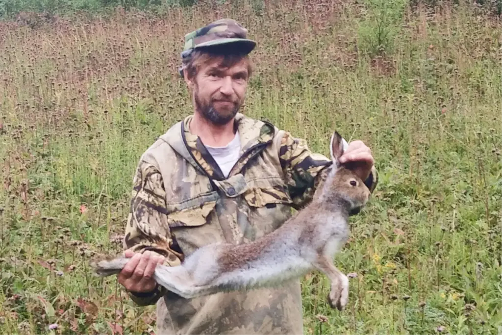 Мужчина в камуфляже держит в руках убитого зайца на фоне поля