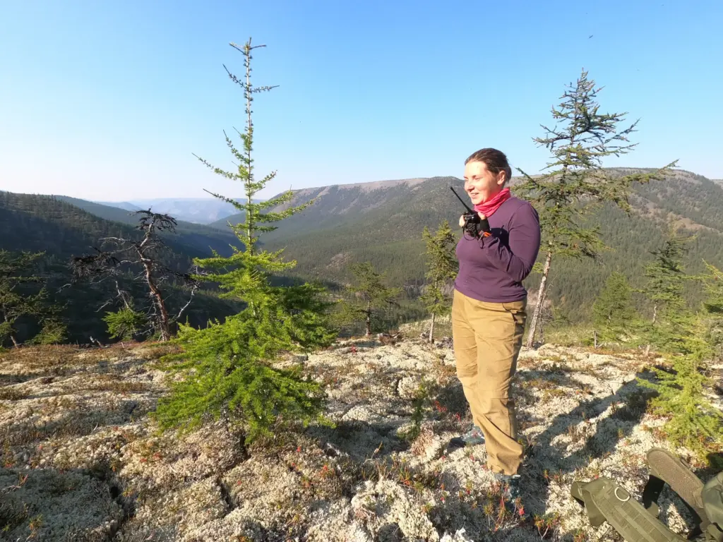 Женщина на горном склоне с рацией в руках даёт команду о завершении электроразведочных работ. На фоне — тайга и хребты Восточной Сибири