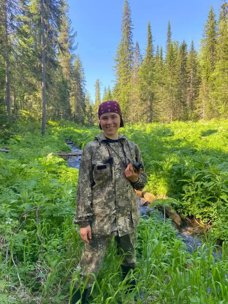 Софья Грудинина, геолог, геохимик.
Женщина в камуфляже стоит в лесу с рацией, на фоне — ручей и густая тайга. Работа по геохимической съёмке в труднодоступной местности