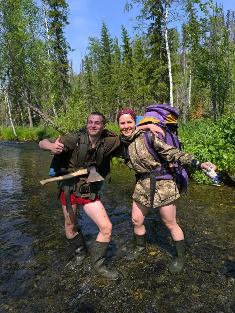 Женщина-геолог с напарником переходит ручей в полевых условиях, одета в сапоги и камуфляж, несёт рюкзак. Оба улыбаются. Настроение бодрое, несмотря на сложный рельеф