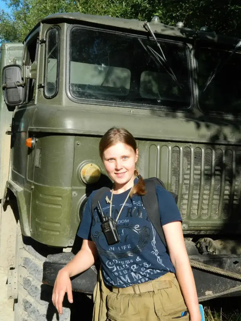 Александра Ис., полевой геолог.
Девушка в повседневной одежде с рацией и рюкзаком стоит у тяжёлой техники (ГАЗ-66). Солнечный день, экспедиционные условия