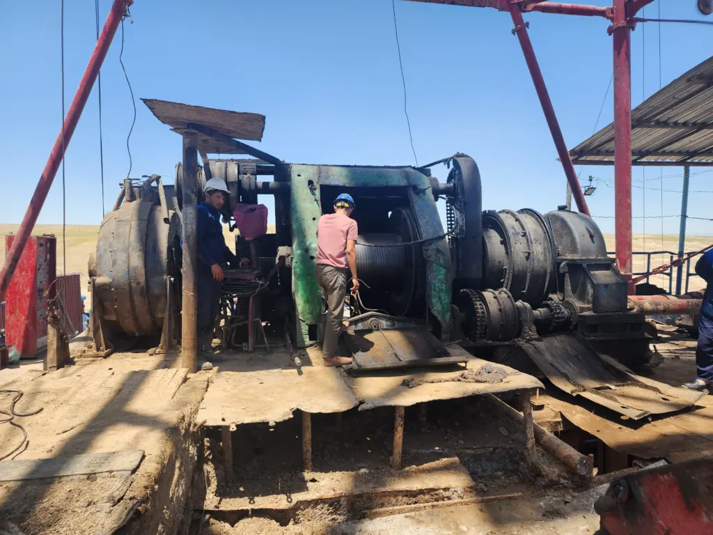 Буровая лебедка на открытой площадке месторождения, бурильщики в касках обслуживают оборудование при эксплуатационном и разведочном бурении скважин на нефть и газ.